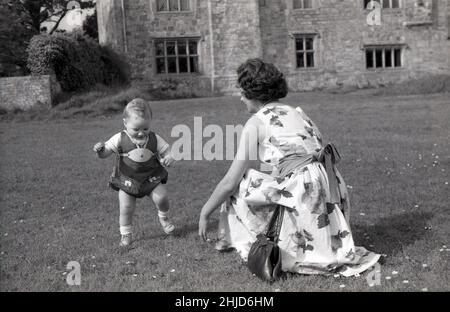 1950s, historical, a mother in a flower patterned dress, common summertime wear in this era, with her infant son, a toddler enjoying perhaps for the first time, freedom from his harness, on a large expanse of lawned area outside a country house, England, UK. Stock Photo