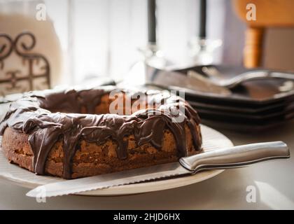 A chocolate bundt cake with fudge glaze waits to be served to guests.  Knife on plate in foreground.  Background includes plates, napkins, forks, cand Stock Photo