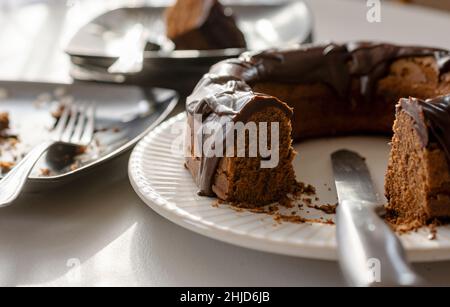 A cut chocolate bundt cake with the knife used sits on white plate.  Black used plate on left blurred in the background with fork.  Far background inc Stock Photo