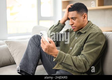 Sad young multiracial man holding smartphone and sitting on the sofa at modern apartment while looking with shocked face and holding his head Stock Photo
