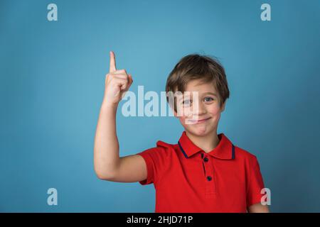 A cute Caucasian boy of 5 years old in a red T-shirt affirmatively and cheerfully points his finger up. Blue background. copy space Stock Photo