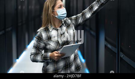 Female data center technician working inside server rack room while wearing face mask Stock Photo