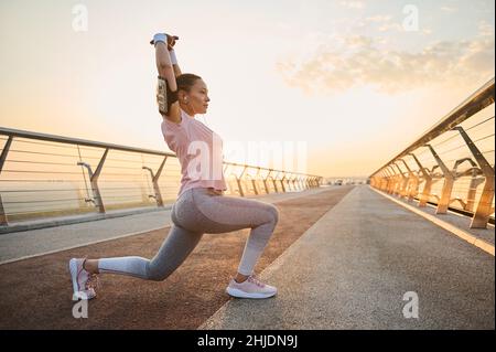 Determined young sportswoman exercising outdoor at dawn. Female athlete in sportswear stretching her arms up, performing squats and lunges, working ou Stock Photo