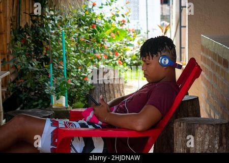 Palomino, Dibulla, La Guajira, Colombia - December 4 2021: Young Man Sitting on the Patio with a Headset and Looking at his Phone Stock Photo