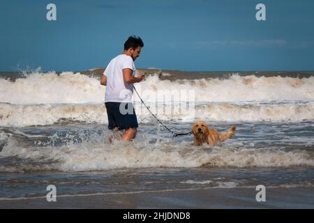 Palomino, Dibulla, La Guajira, Colombia - December 4 2021: Young Latin Man Talking on the Phone and Walking his Leashed Dog in the Sea Stock Photo