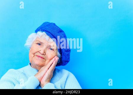 beautiful retired woman in blue beret and warm pullover looking at camera and smile in blue wall background in studio Stock Photo