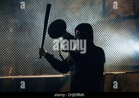 Angry masked man with a bat is shouting into the megaphone. Stock Photo