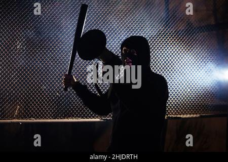 Angry masked man with a bat is shouting into the megaphone. Stock Photo