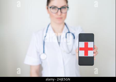 Female doctor in glasses holds a smartphone with a red cross on the screen. Serious nurse in a medical coat with a stethoscope on his shoulders Stock Photo