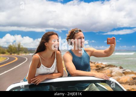 Road trip vacation sports car people taking phone pictures of Hawaii landscape on summer travel trip. Tourists couple taking photos with smartphone Stock Photo