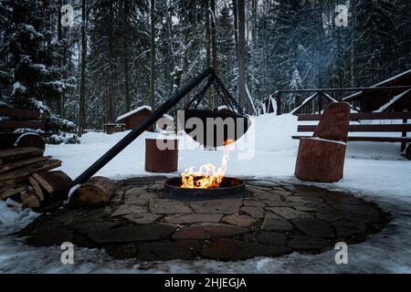 Over the fire hangs a pot in which to cook food. On a hook on a tripod, steam comes out of the pan. Winter Camping outdoor cooking Stock Photo