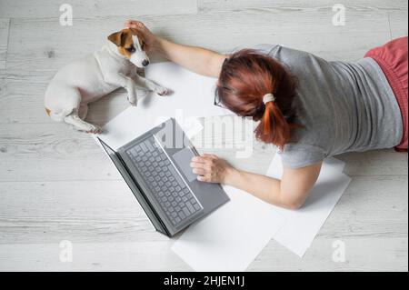 A woman lies on her stomach on the floor and is typing on a laptop. Girl freelancer works remotely from home with a puppy. Jack Russell Terrier next Stock Photo