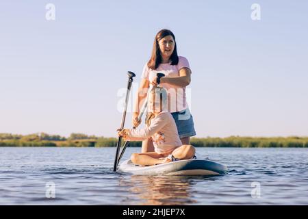 Woman of middle age and daughter rowing sitting on sup board with great efforts, exercising and enjoying time with green reeds and trees in background Stock Photo
