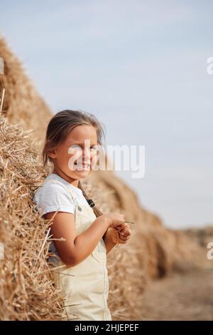 Blissful young female child standing near high haymow in field with braid on shoulder, hands holding hay with squinting eyes looking somewhere far Stock Photo