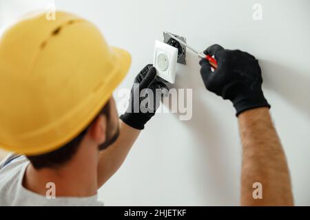 Professional electrician in uniform using screwdriver while installing electrical socket outlet after renovation work Stock Photo