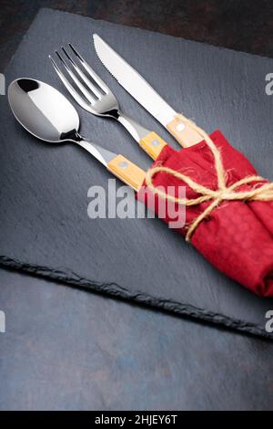 Cutlery with in a burgundy napkin on a dark background. Spoon, fork and knife on a slate board. Table setting. Top view Stock Photo