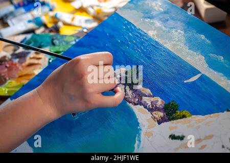 Artist's hand with a brush, the process of drawing oil paintings with the sea. Tubes of paint on a blurred background. Selective focus. Stock Photo