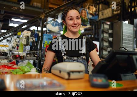 Positive young female worker in casual clothes with tattoo on arm and creative hairstyle smiling happily and looking at camera standing at cash regist Stock Photo