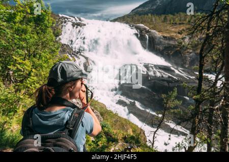 Kinsarvik, Hordaland, Norway. Young Woman Lady Tourist Traveler Taking Pictures Of Waterfall Nyastolfossen In Hardangervidda Mountain Plateau Stock Photo
