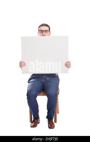 Businessman seated on chair holding a big blank banner with copy space for messages. Full length business person with empty advertising placard. Warni Stock Photo