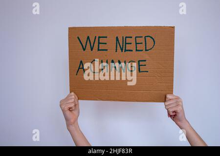 Hand holding cardboard with 'we need a change' inscription over gray wall background. She may be protesting global warming or environmental pollution. Stock Photo