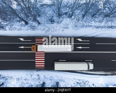 Two white trucks on the road go in different directions. Dangerous red and white pedestrian crossing on the highway. Top view of the road in winter Stock Photo