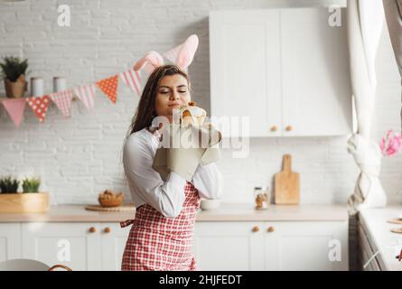 A beautiful young girl in rabbit ears holds an Easter cake in her hands, sniffs a pleasantly smelling freshly baked cake stands near the table in the Stock Photo