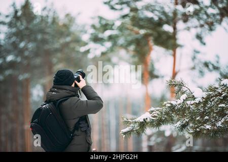 Young Man Backpacker With Photo Camera Taking Photo In Winter Snowy Forest. Active Hobby. Hiker Walking In Snowy Pinewood Forest Stock Photo