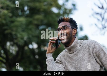 Close up of smiling African American man in turtleneck vest sitting on park bench and talking on his mobile phone Stock Photo