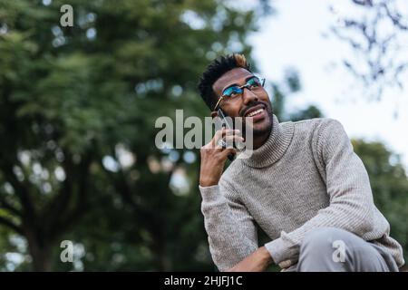 Portrait of smiling African American man in turtleneck vest sitting on park bench and talking on his mobile phone Stock Photo