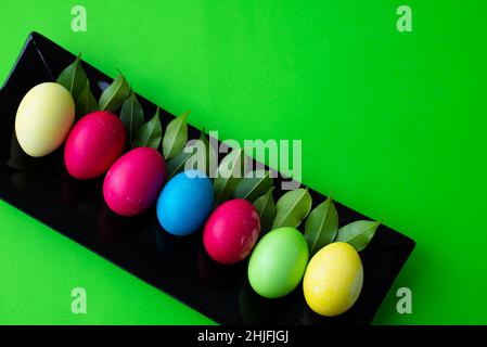 Easter templates for designs. Corner for text. On a rectangular black dish lies a row of multi-colored painted eggs and a row of green leaves. Stock Photo