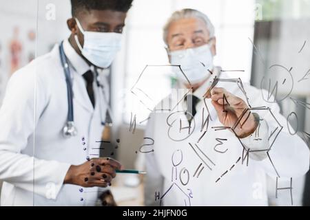 African american and caucasian scientists in white lab coats and masks writing various formulas on flip chart. Two competent doctors checking and examining together new virus. Stock Photo