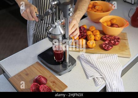 glass of freshly squeezed pomegranate juice, chopped pomegranates and a pomegranate juicer at home. A man in a striped dark apron squeezes juice from Stock Photo