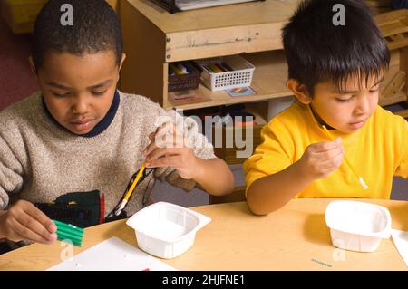 Education Preschool classroom 4-5 year olds art activity two boys sitting side by side using paint brushes to apply glue, using opposite hands Stock Photo
