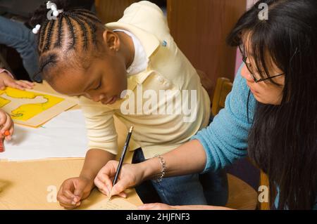 Education Preschool classroom 4-5 year olds female teacher with girl writing down what the girl wants to say about her drawing Stock Photo