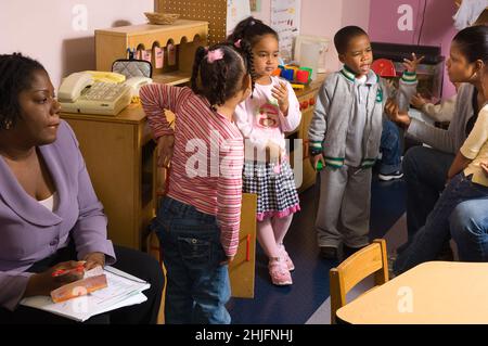 Education Preschool classroom ages 4-5 observer watches as female teacher works with two children to resolve dispute, two other children waiting to st Stock Photo