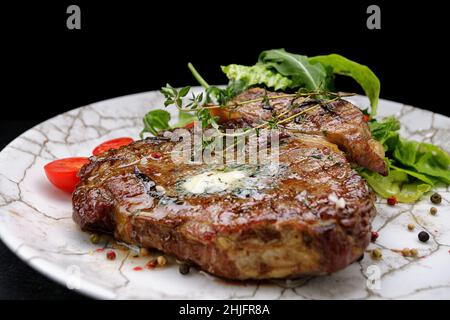Grilled meat steak with butter, herbs and cherry tomatoes on a white plate, against a dark background Stock Photo