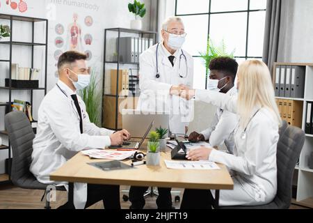 Group of four multiracial doctors in face masks greeting each other with fist bump during coronavirus pandemic. Medical workers sitting at desk with modern gadgets and having meeting. Stock Photo