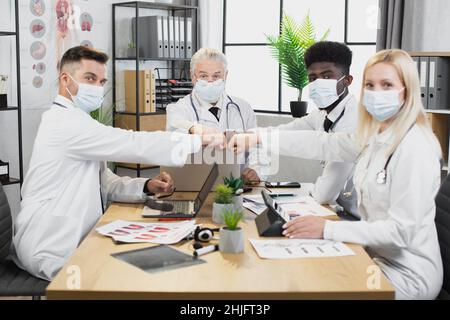 Fout multicultural medical experts in face masks and lab coats greeting with fists on meeting. Team of doctors sitting at table with various modern gadgets and looking at camera. Stock Photo