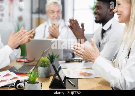 Close up of multicultural medical workers applauding after successful conference at hospital room. Team of doctors sitting at desk with modern technologies. Stock Photo