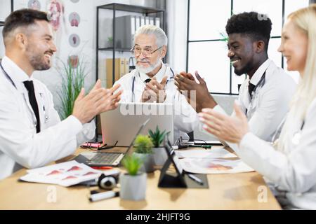 Group of multiethnic healthcare experts clapping in hands during medical conference. Happy colleagues smiling to each other while sitting at desk with various modern gadgets. Stock Photo