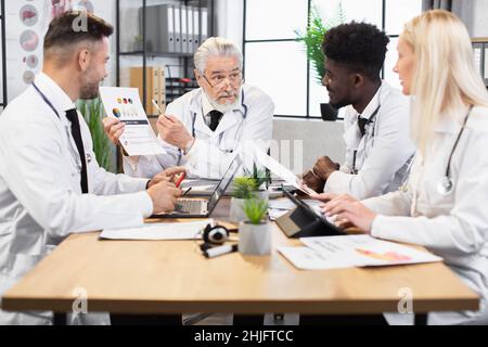 Multiracial professional biochemists in white lab coats discussing dna formula during conference. Modern gadgets for international meeting. Stock Photo