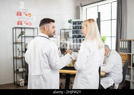 Caucasian male and female medical scientists in white lab coats having discussion while standing near glass flip chart at hospital room. Researchers, cooperation and brainstorming concept. Stock Photo
