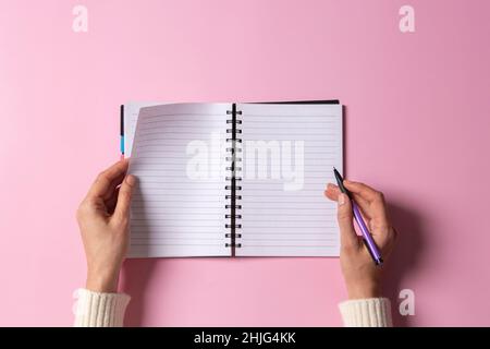 Female hand writing in blank open spiral notebook with pen on pastel pink background. Top view, copy space. Education, remote work, business, finance Stock Photo