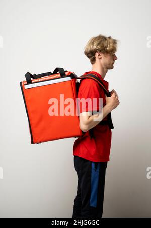 Fast Food Delivery Service Concept. Side view of smiling male courier wearing red uniform and holding thermal backpack bag isolated on studio Stock Photo