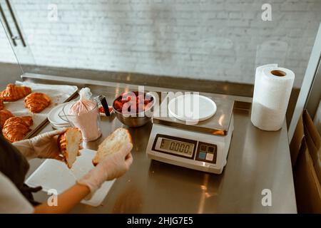 Confectioner holds cut croissant near metal table with ingredients and scales in bakery Stock Photo
