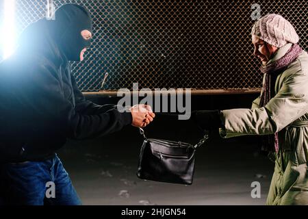Crime scene. Robber in mask trying to snatch bag from young woman. Stock Photo