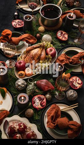 Christmas holiday dinner festive table setting with roast chicken, fruit, poached pears, hot spiced wine. Black linen table with dishes, candle, baubl Stock Photo