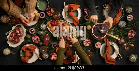 Christmas or New Year holiday dinner festive table setting. Flat-lay of human hands feasting over dark table with chicken, fruit, hot spiced wine, dis Stock Photo