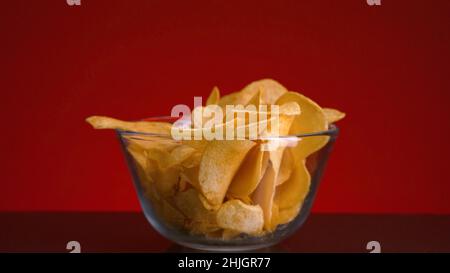 Close-up of hand picking up chips. Stock footage. Person takes one potato chip from bowl on isolated background. Chips in transparent bowl on red back Stock Photo